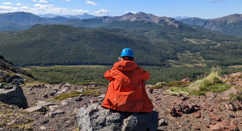 A person wearing a helmet sits with their back facing the camera. They are looking out over a vast mountainous landscape. 